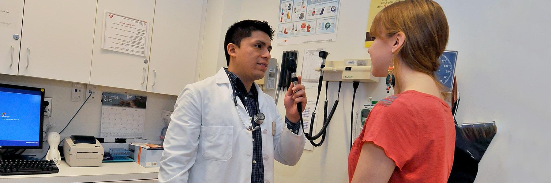 UMass Boston staff member in white coat with stethoscope examines student sitting on a medical table