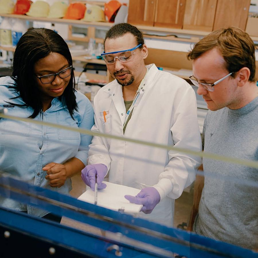 A grad student wearing a lab coat teaches 2 students in an environmental lab.